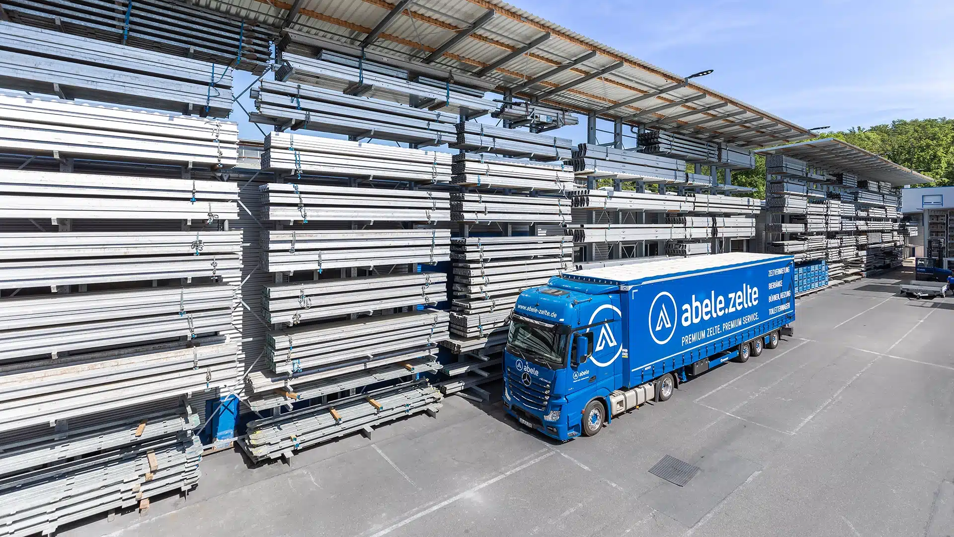 Unternehmen 14 Blauer Lastwagen mit Firmenlogo geparkt vor Stapeln von Aluminiumprofilen unter überdachter Lagerhalle bei sonnigem Wetter.