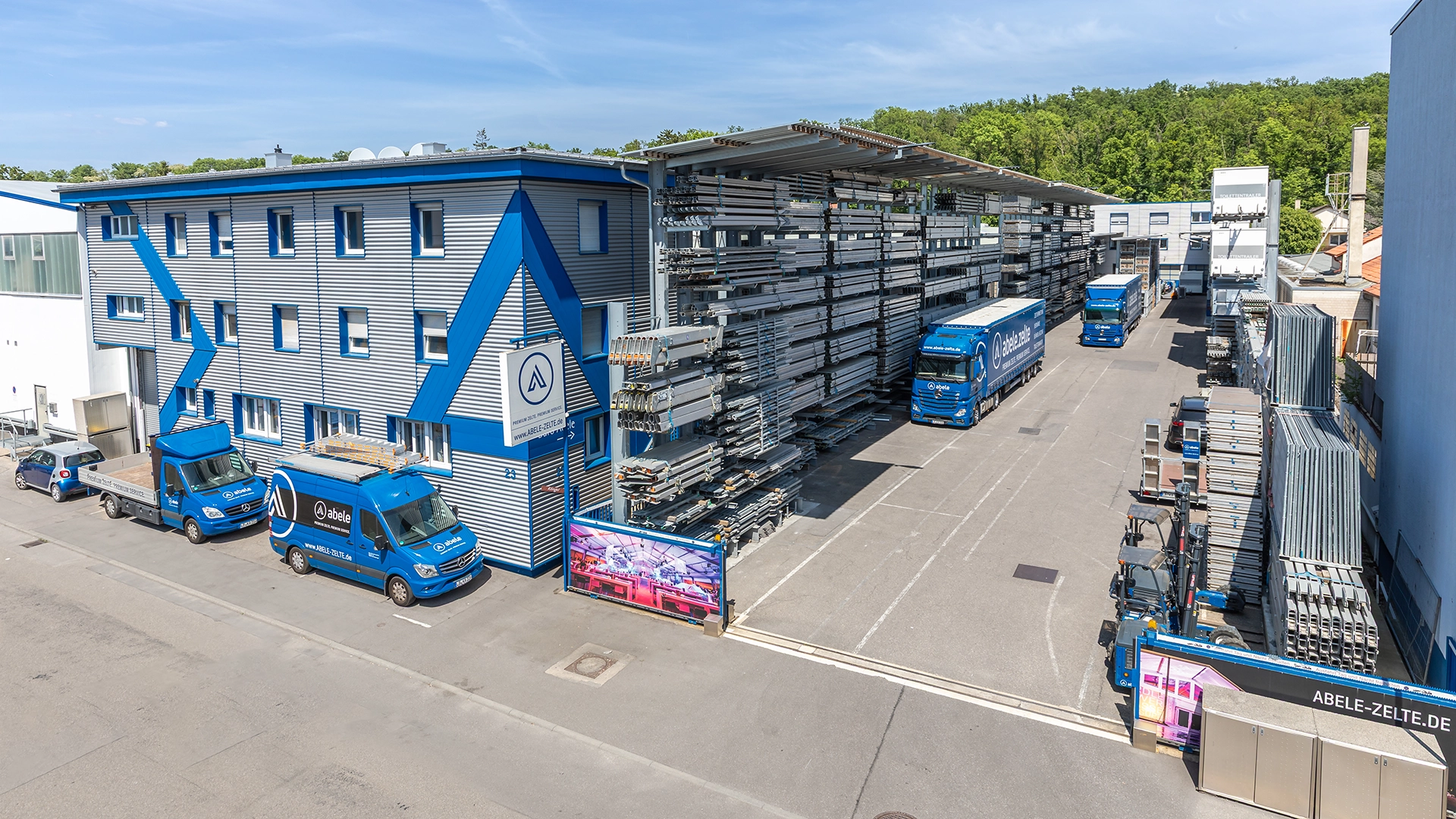 Unternehmen 13 Industriegebäude mit blauen Fahrzeugen und großen Regalstrukturen im Hof, umgeben von grünen Bäumen im Hintergrund.