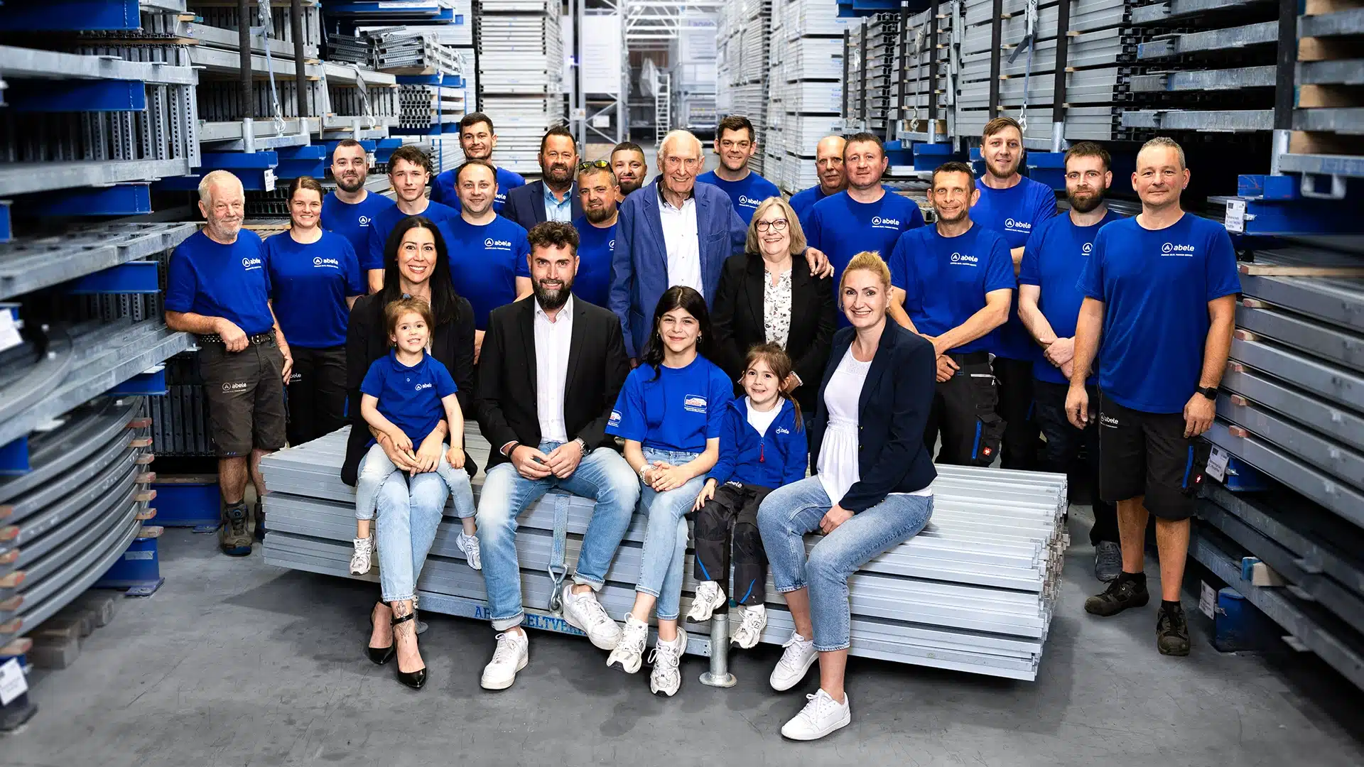 Unternehmen 12 Gruppe von 22 Menschen in blauer Arbeitskleidung in einem Lager, posieren lächelnd vor gestapelten Metallregalen.