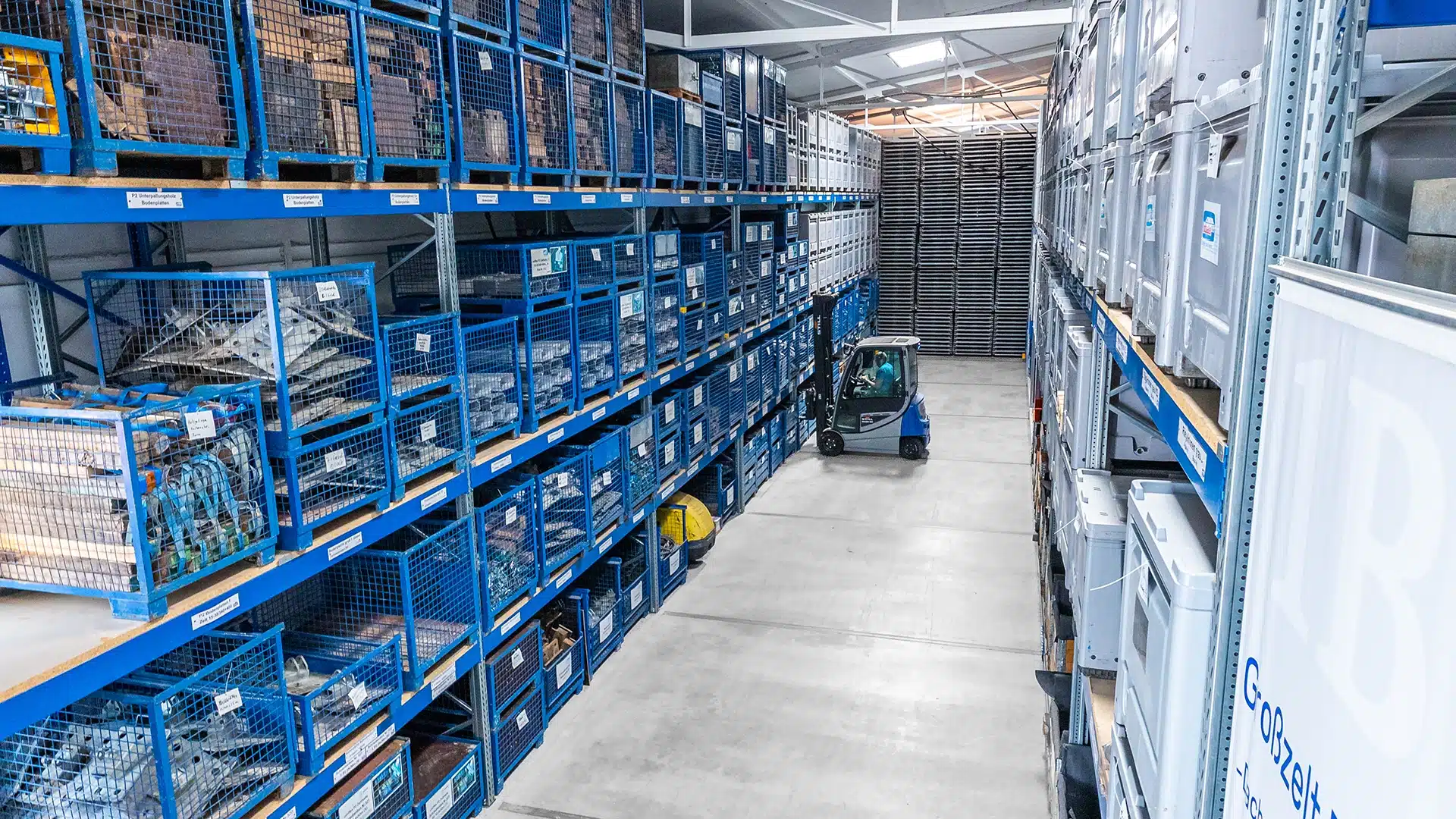 Unternehmen 10 Lager mit blauen Regalen voller Kisten und einem kleinen Gabelstapler auf einem breiten Gang, von oben betrachtet.