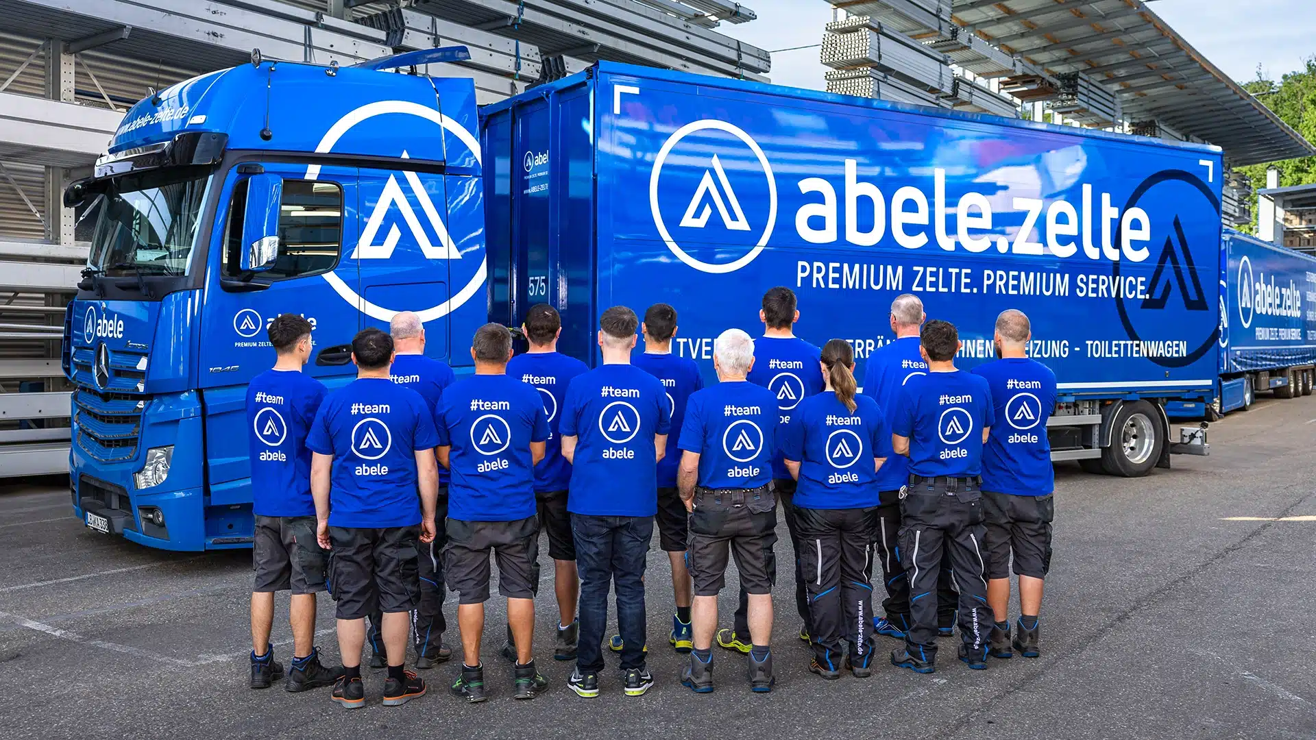 Unternehmen 7 Gruppe von Mitarbeitern in blauen T-Shirts steht vor einem blauen Abele-Lastwagen mit Firmenlogo und Beschriftung.