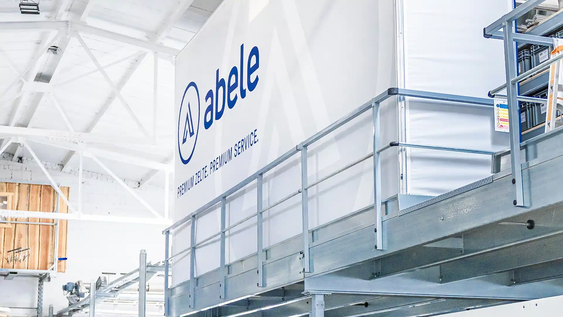 Unternehmen 6 Außenansicht eines großen, weißen Lagergebäudes mit abele-Logo und Slogan „Premium Zelte, Premium Service“ in blauen Buchstaben.