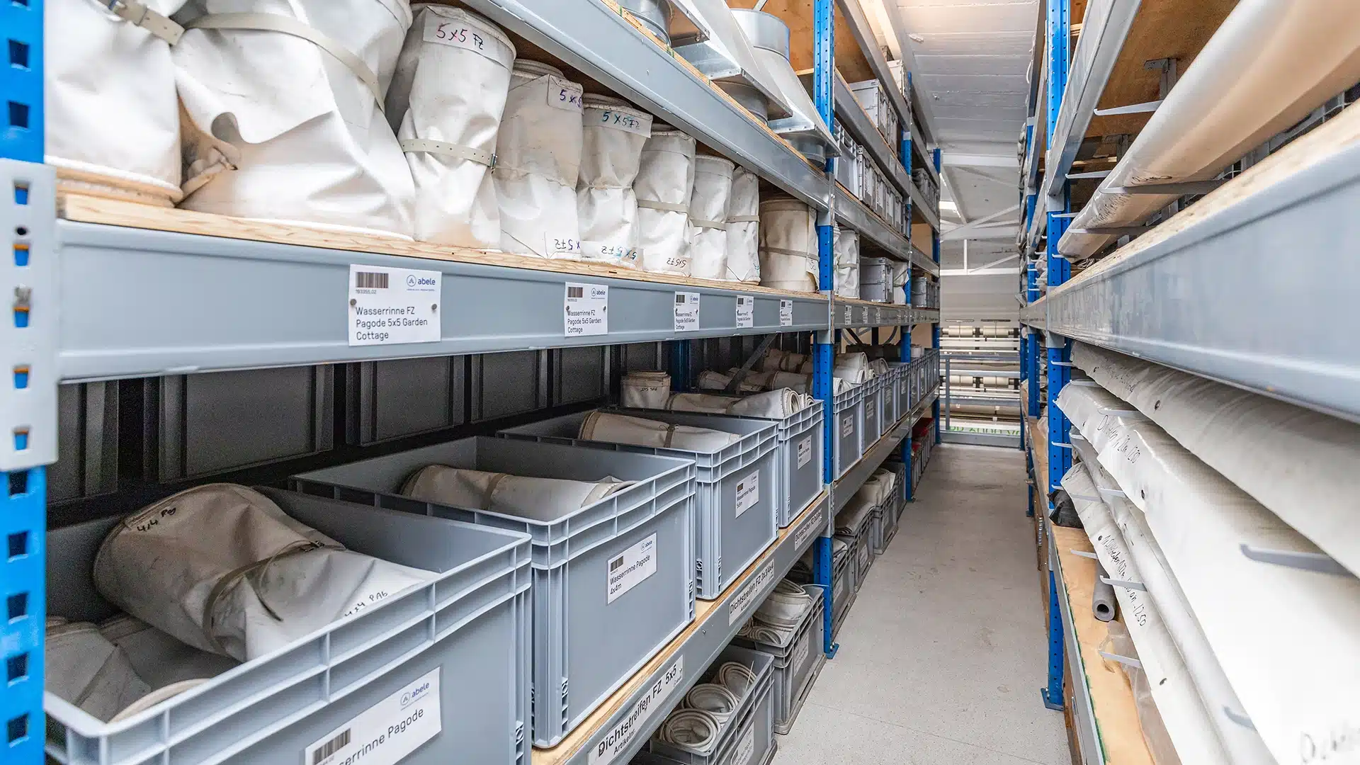 Unternehmen 4 Lagerregal mit grauen Boxen, beschriftet mit „Wasserrinne“ und weißer Plane, in einem gut organisierten Lagerraum.