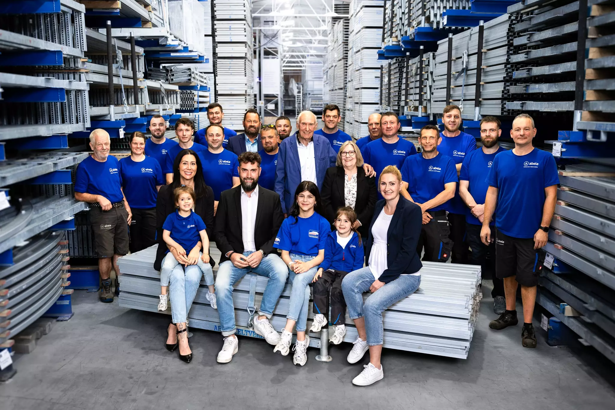 Gruppe von 20 Menschen in blauer Arbeitskleidung posiert in einem Lager mit Metallregalen und Stapeln.