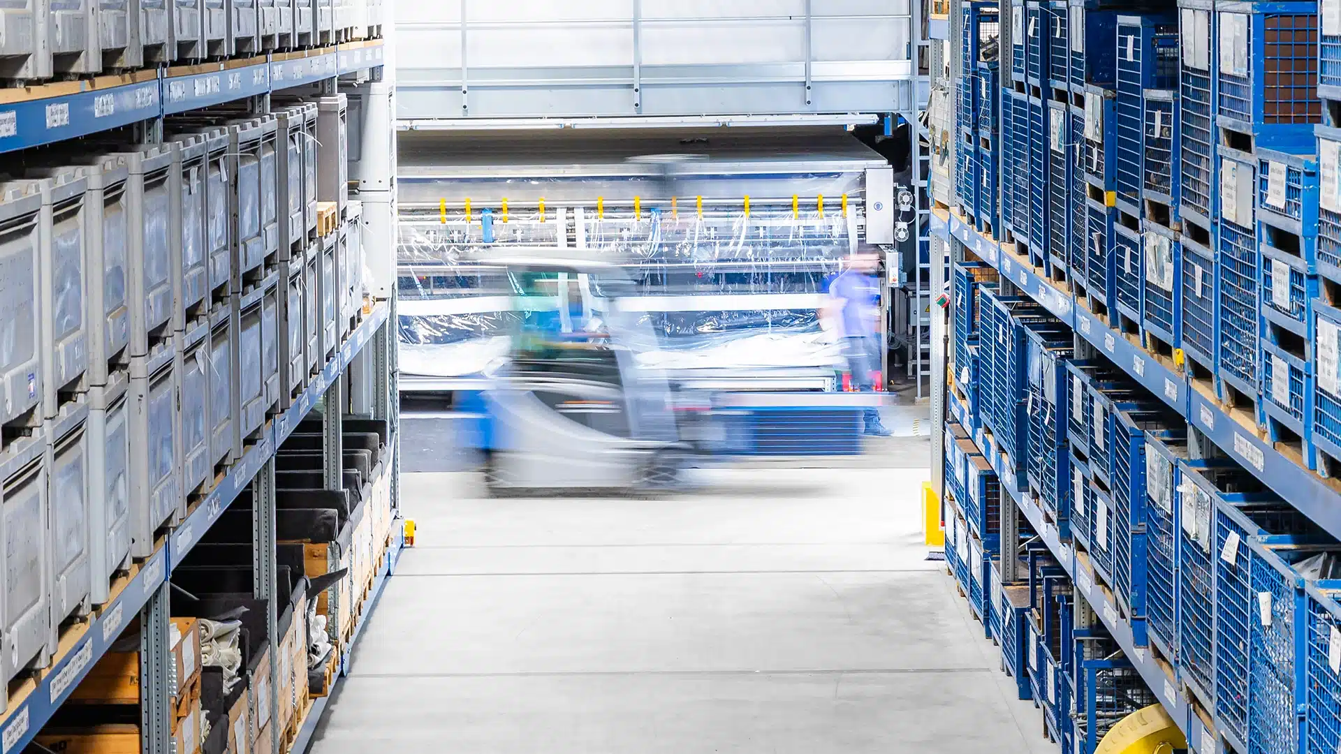 Unternehmen 11 Gabelstapler fährt schnell durch ein großes Lager mit hohen Regalen voller blauer und grauer Kisten.
